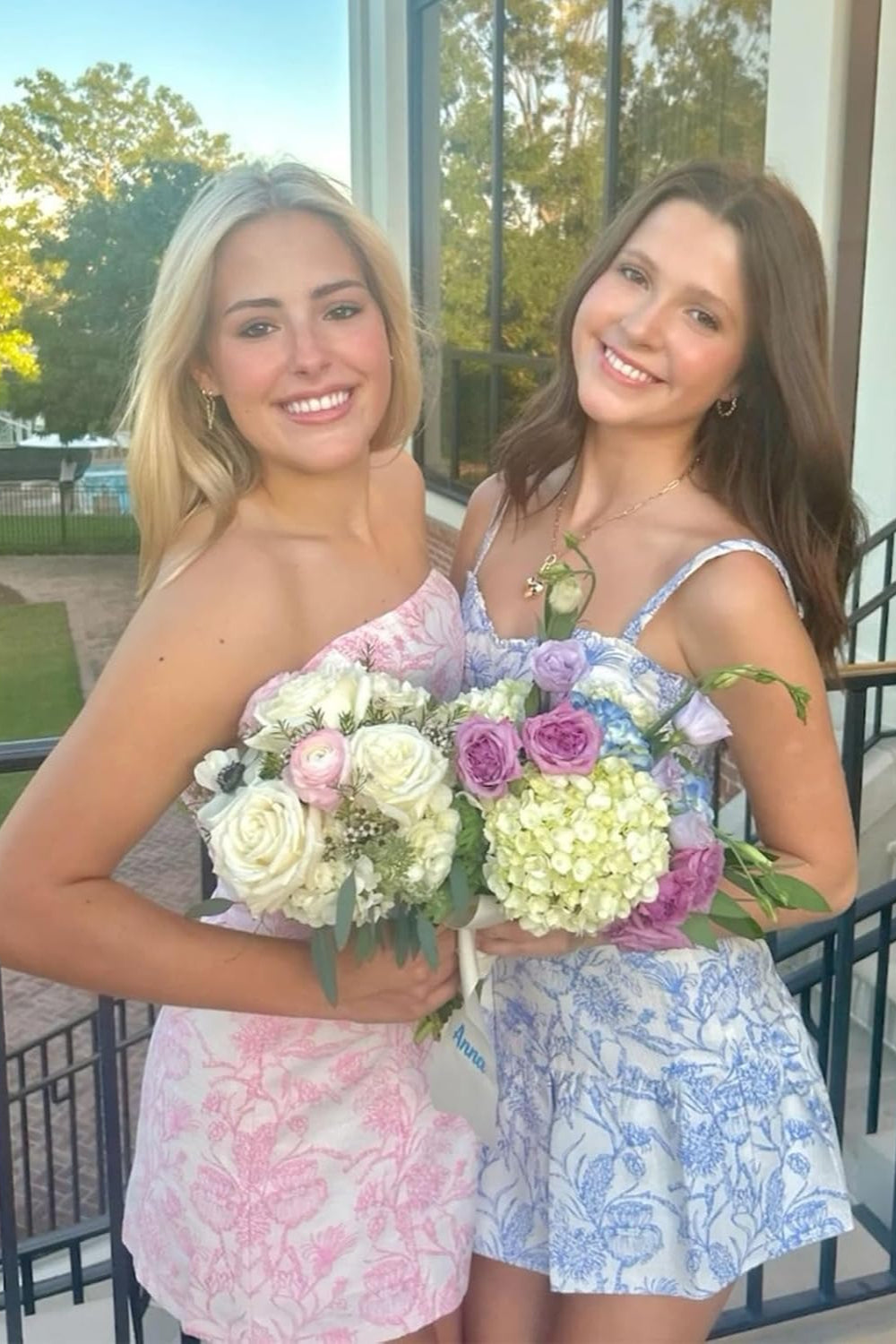 Two smiling models in 27dress Floral Homecoming Dresses pastel pink and blue floral dresses holding bouquets on balcony